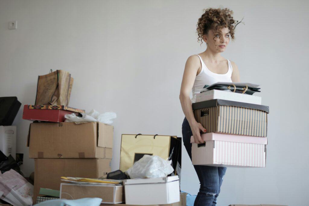 A woman is carrying a carton and is surrounded by items for packing.